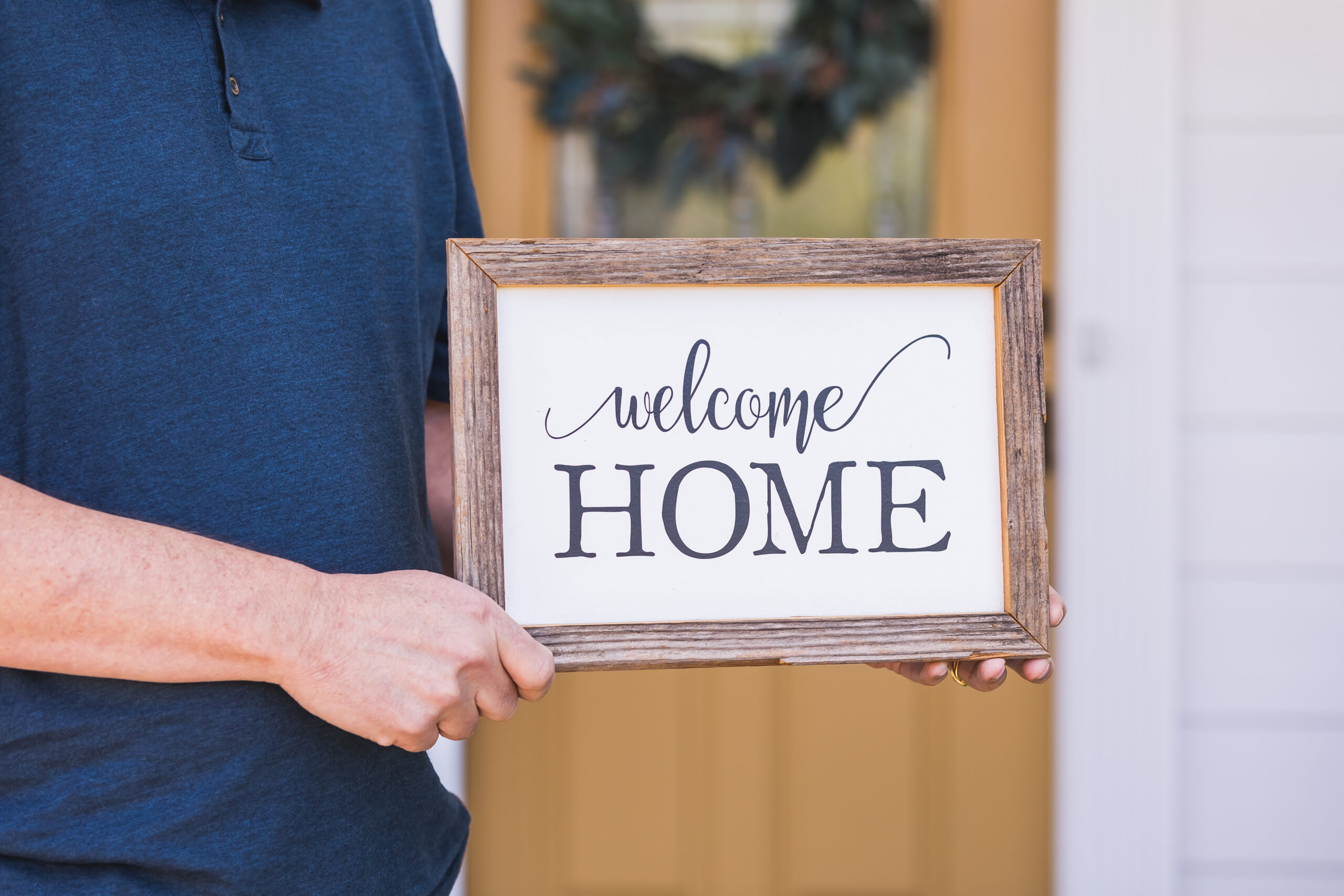 Brand photography photo by Ann Keen Photography. Close up photo of a man holding a Welcome Home sign in front of a house. Headshot and Brand Photography
